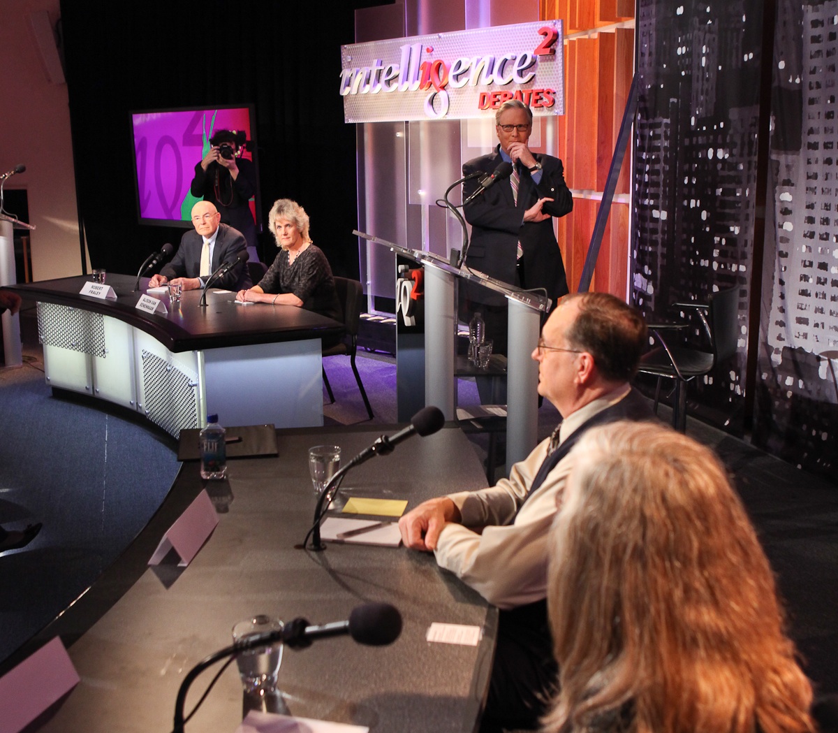 photo of a stage showing a moderator with two tables of two debaters each