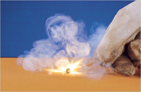 A person wearing white work gloves striking a match on a light brown surface against a blue background.