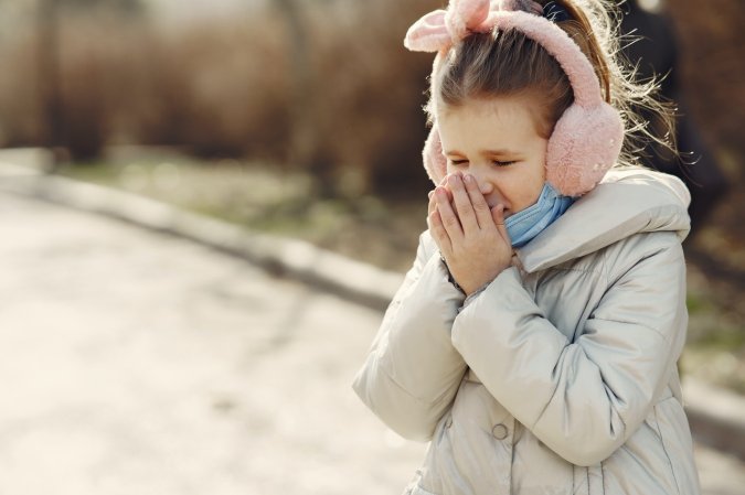 A young girl stands outside in a winter coat, pink earmuffs, and a blue face mask pulled down over her chin as she sneezes into her hands.