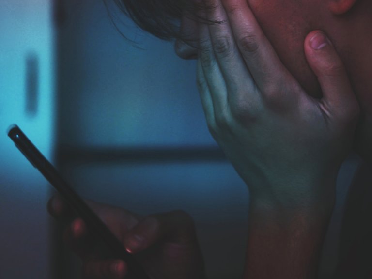 A person with their hand on their face while they look at their phone in a dimly lit room.