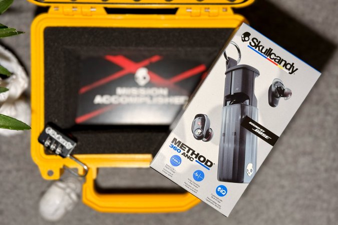 Black Skullcandy Method ANC earbuds retail product sitting on top of a yellow pelican case on a grey felt background